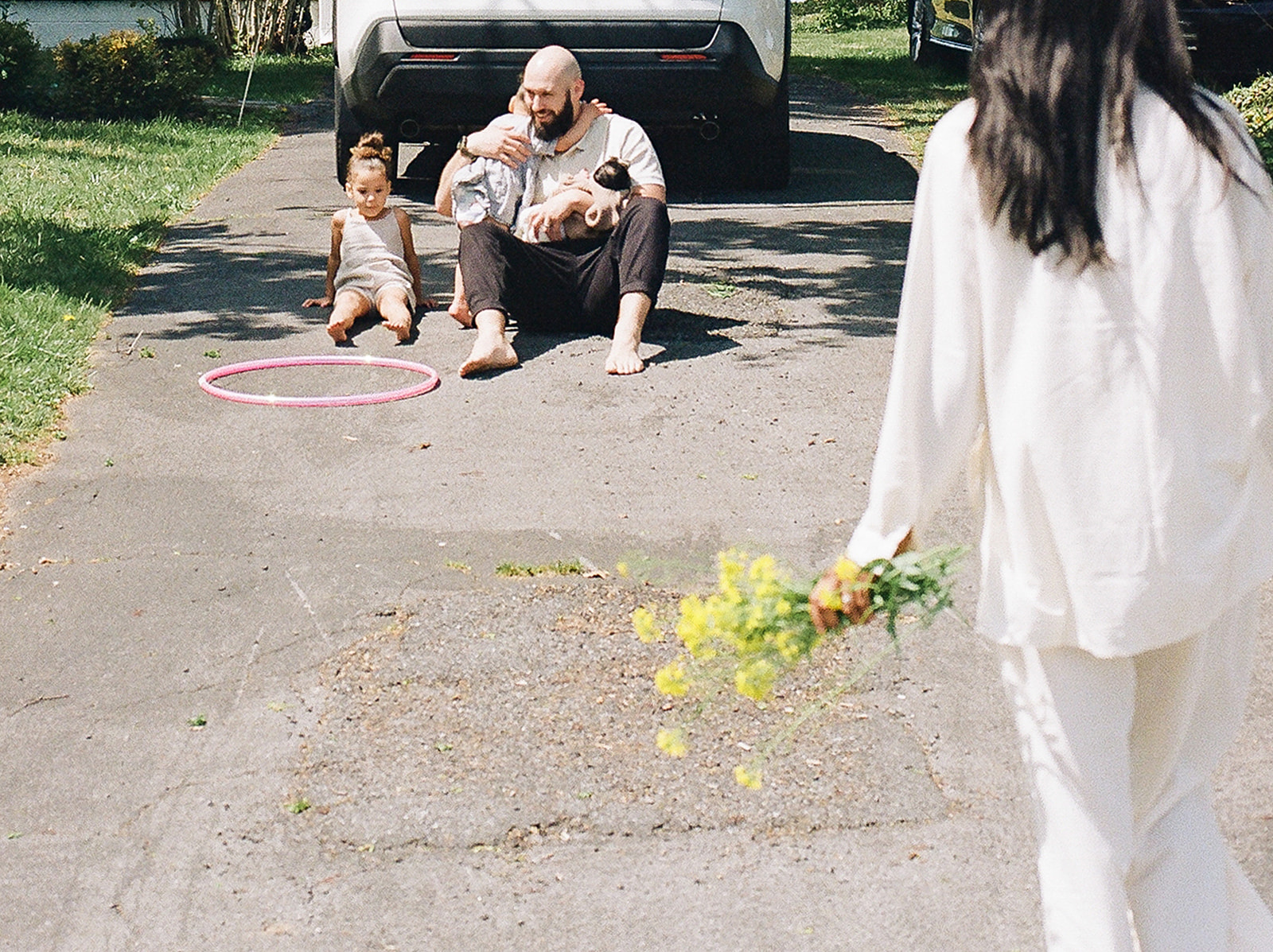 Film family photos of a woman with a bouquet of flowers walking towards her husband and children 