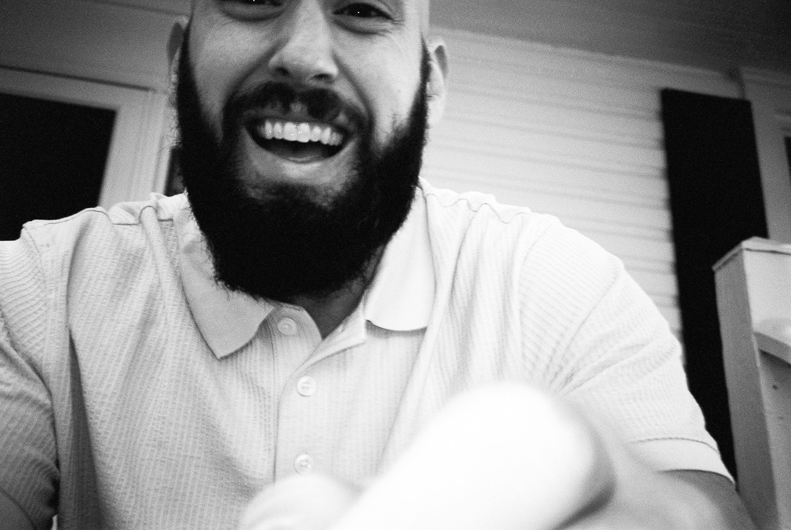 A black and white film photo of a man with a thick beard smiling as he reaches for the camera 