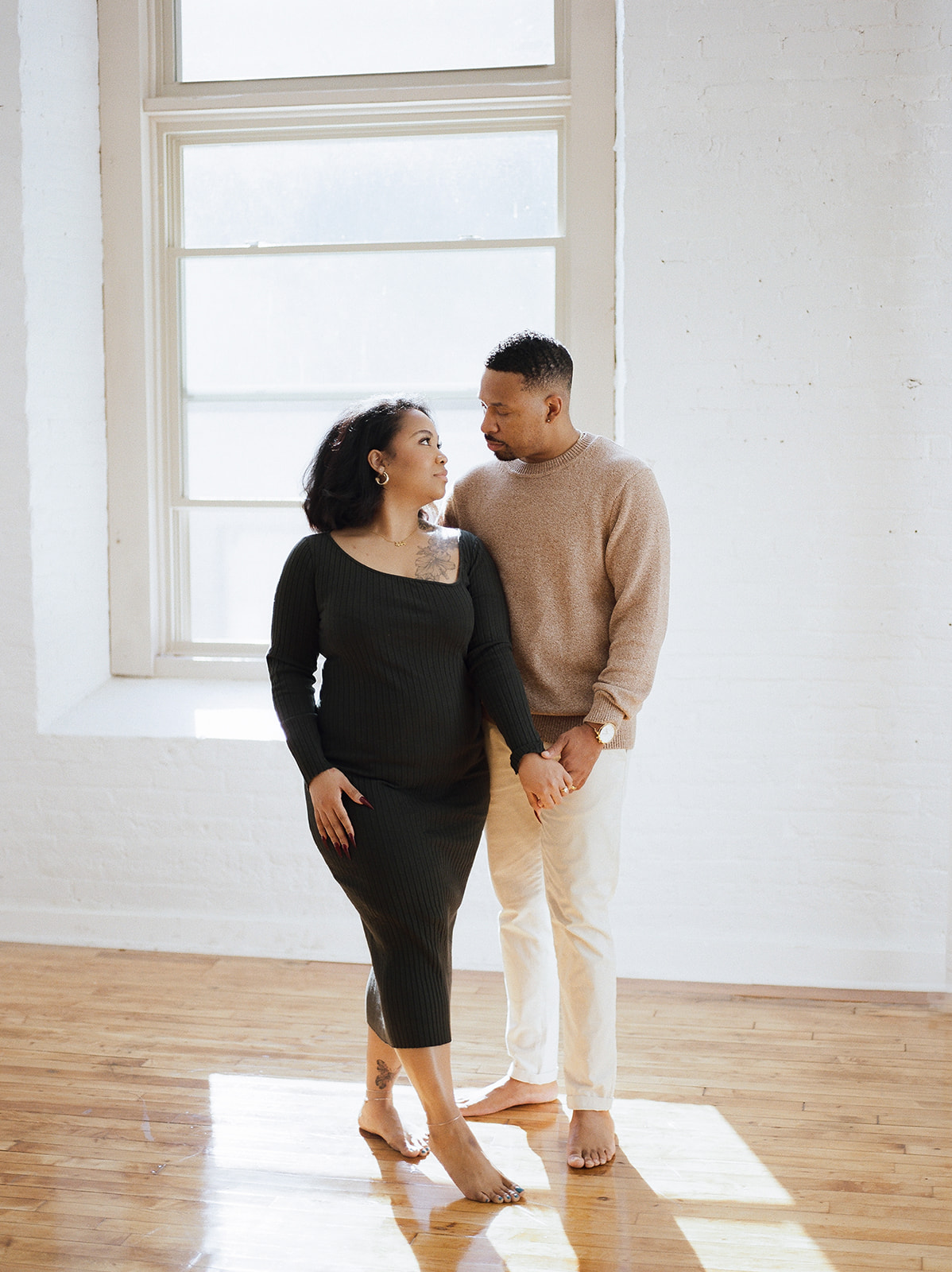 A pregnant woman and her husband pose for studio family photos in a patch of light on a hardwood floor