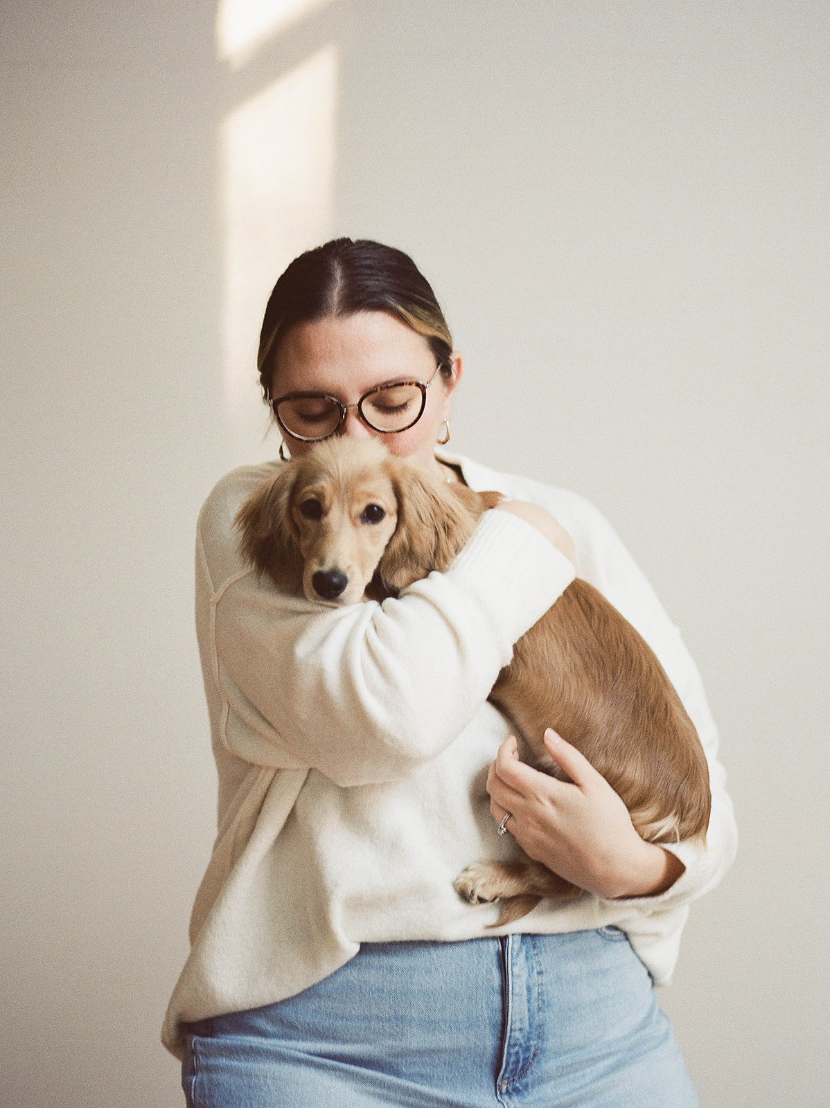 A woman with glasses wearing a white sweater and jeans sits on a stool holding a dachshund