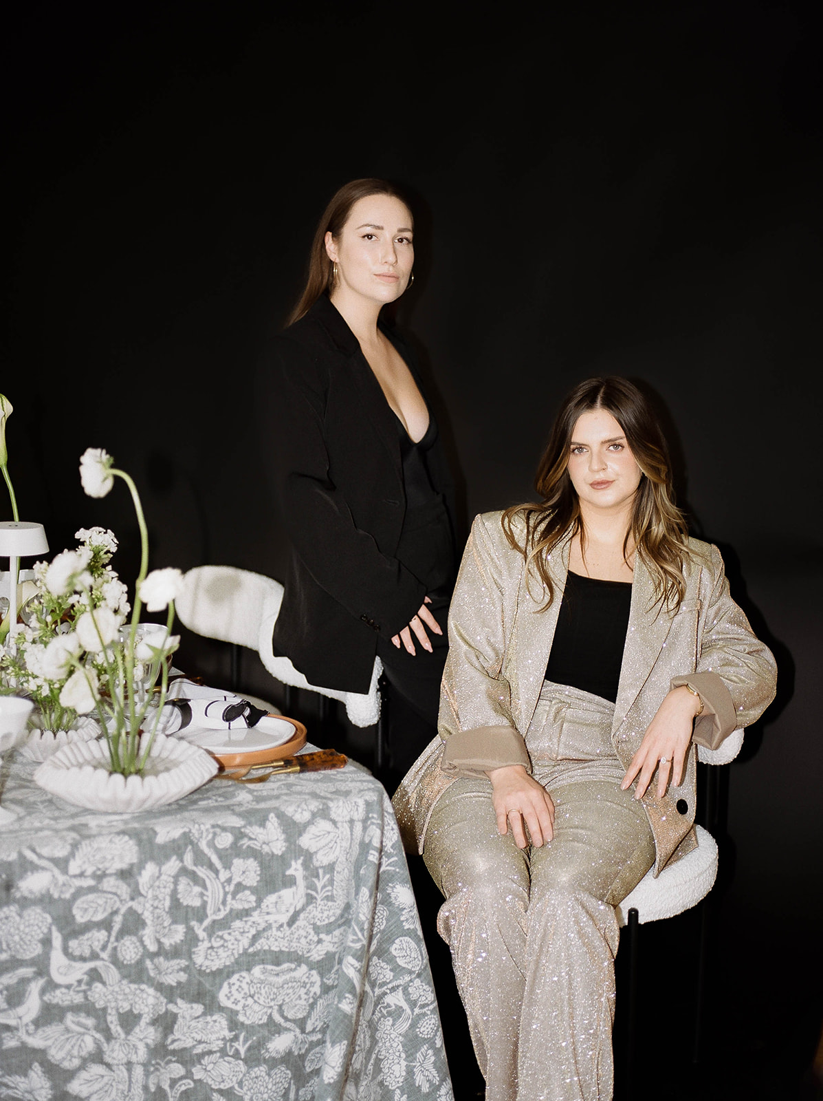 Two women looking at the camera in suits, one sitting one standing by a table with flowers and place setting