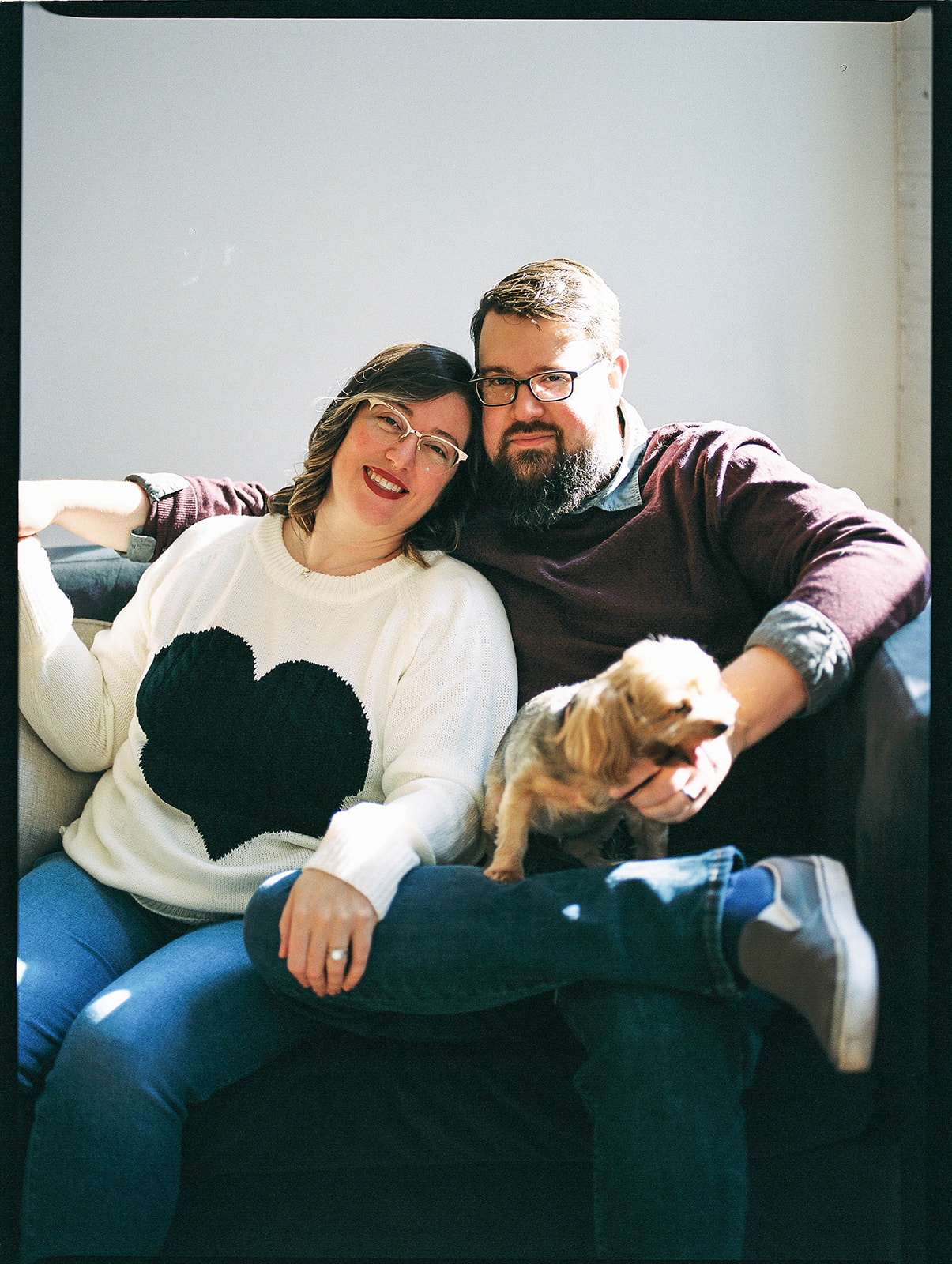 A husband and wife sit on a sofa with a small dog