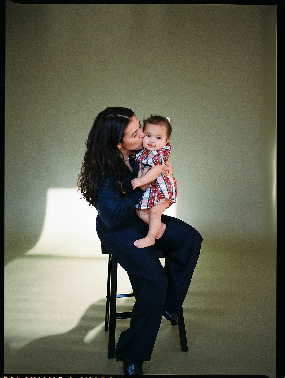 A mother sits on a stool and kisses her baby in these studio family photos