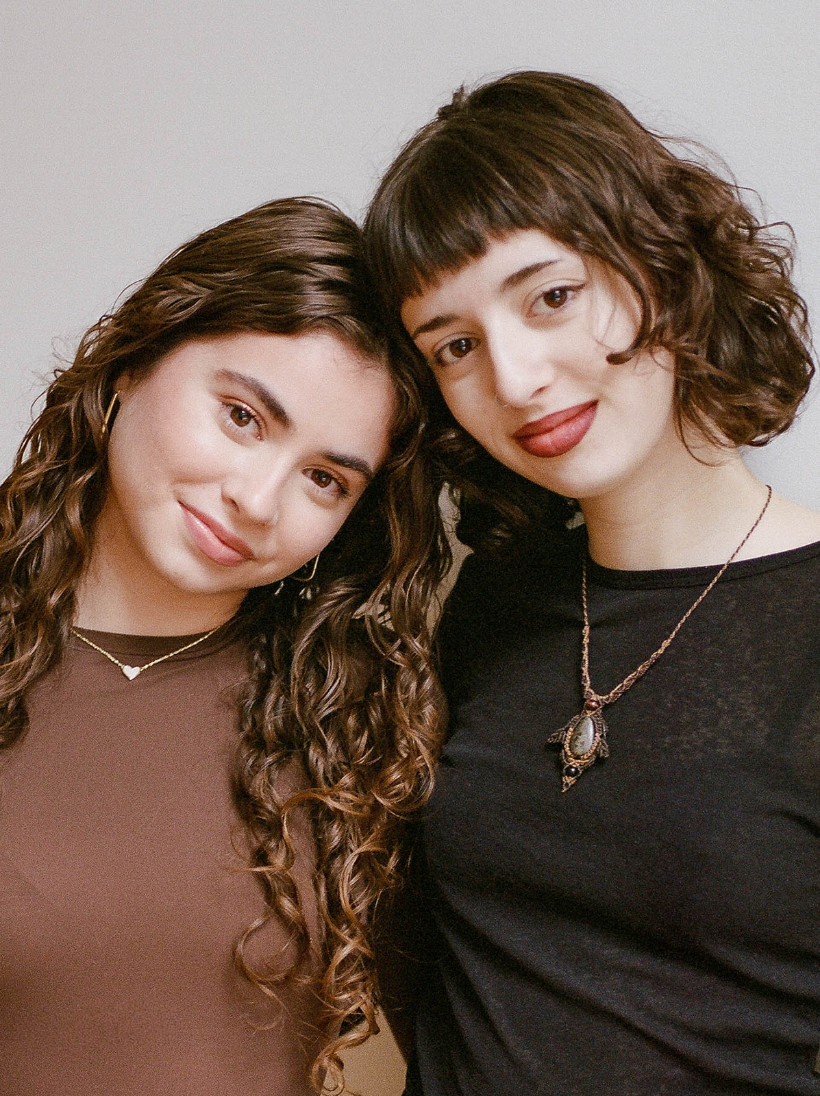 Two sisters with brown curly hair and large brown eyes look at the camera with their heads tilted together