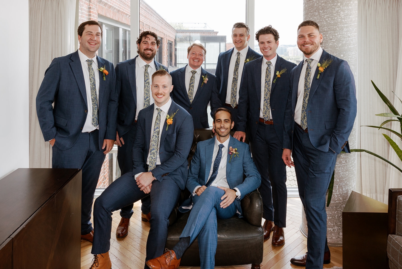 A groom sits in a leather chair surrounded by his groomsmen, wearing blue suits and floral ties