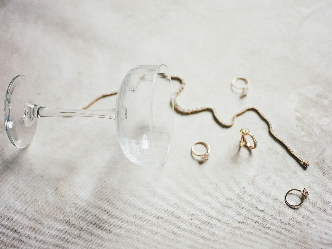 A champagne glass on it's side that looks like it has spilled a gold necklace and four gold rings onto a white tablecloth