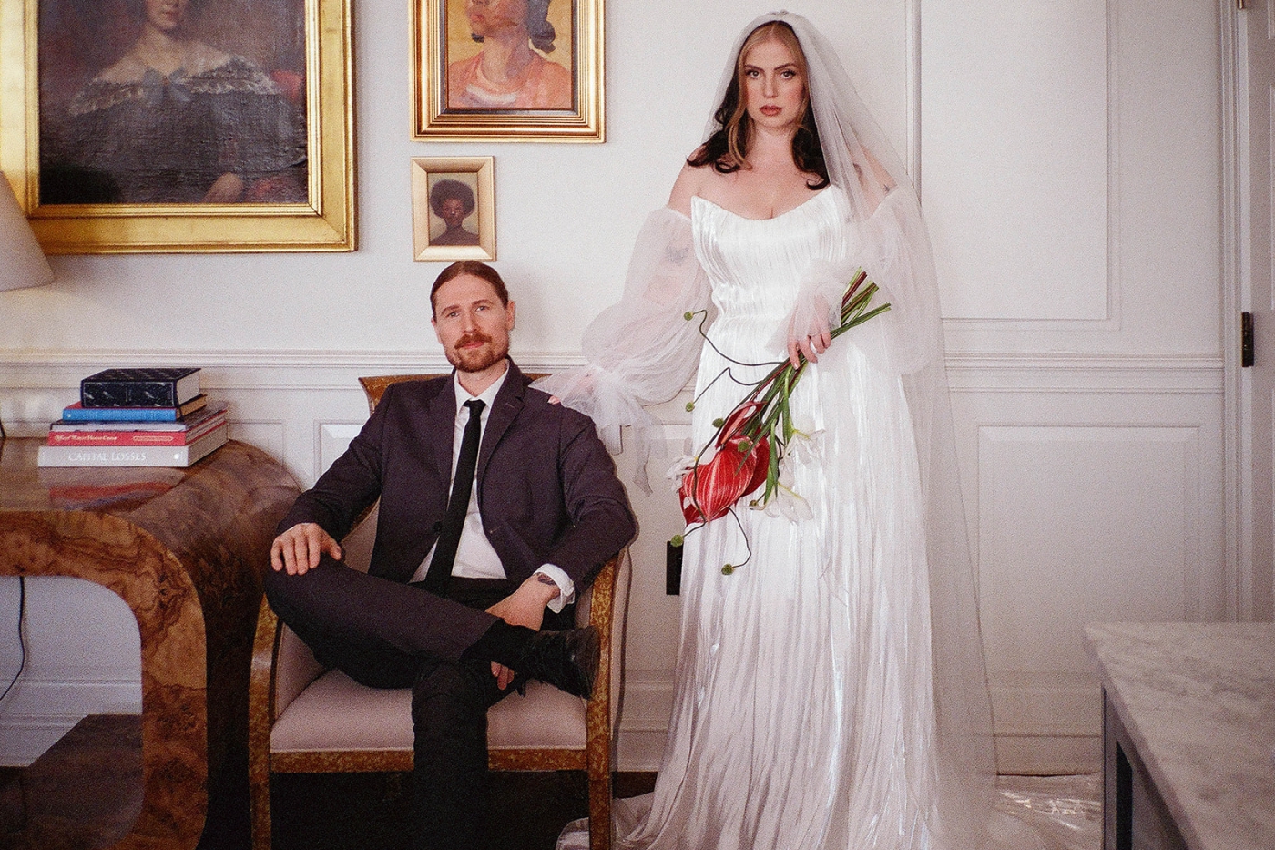 A groom sits in a chair while a bride stands beside him holding a bouquet