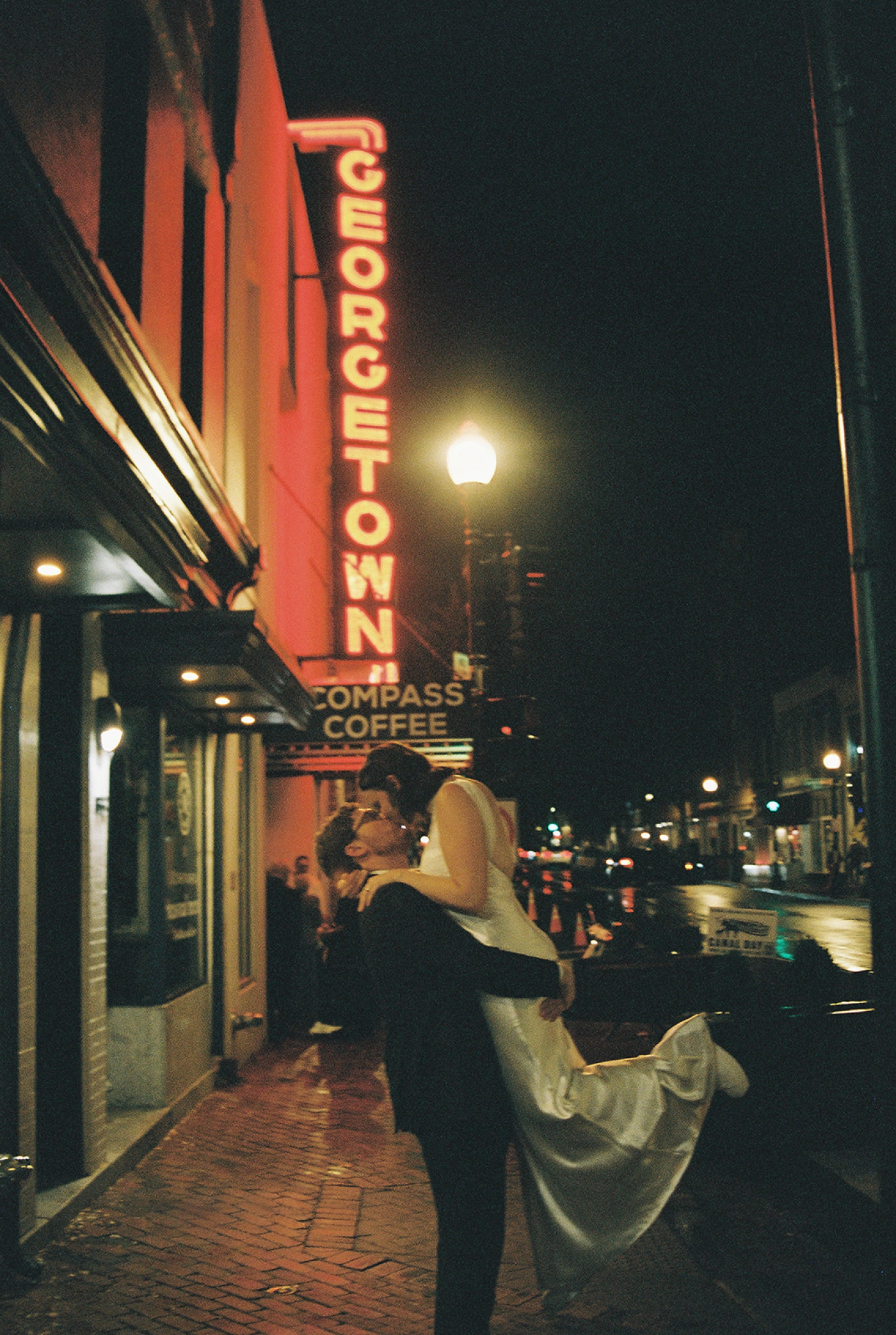 A groom lifts the bride into the air and kisses her in front of a neon Georgetown sign outside of the Fathom Gallery in DC 