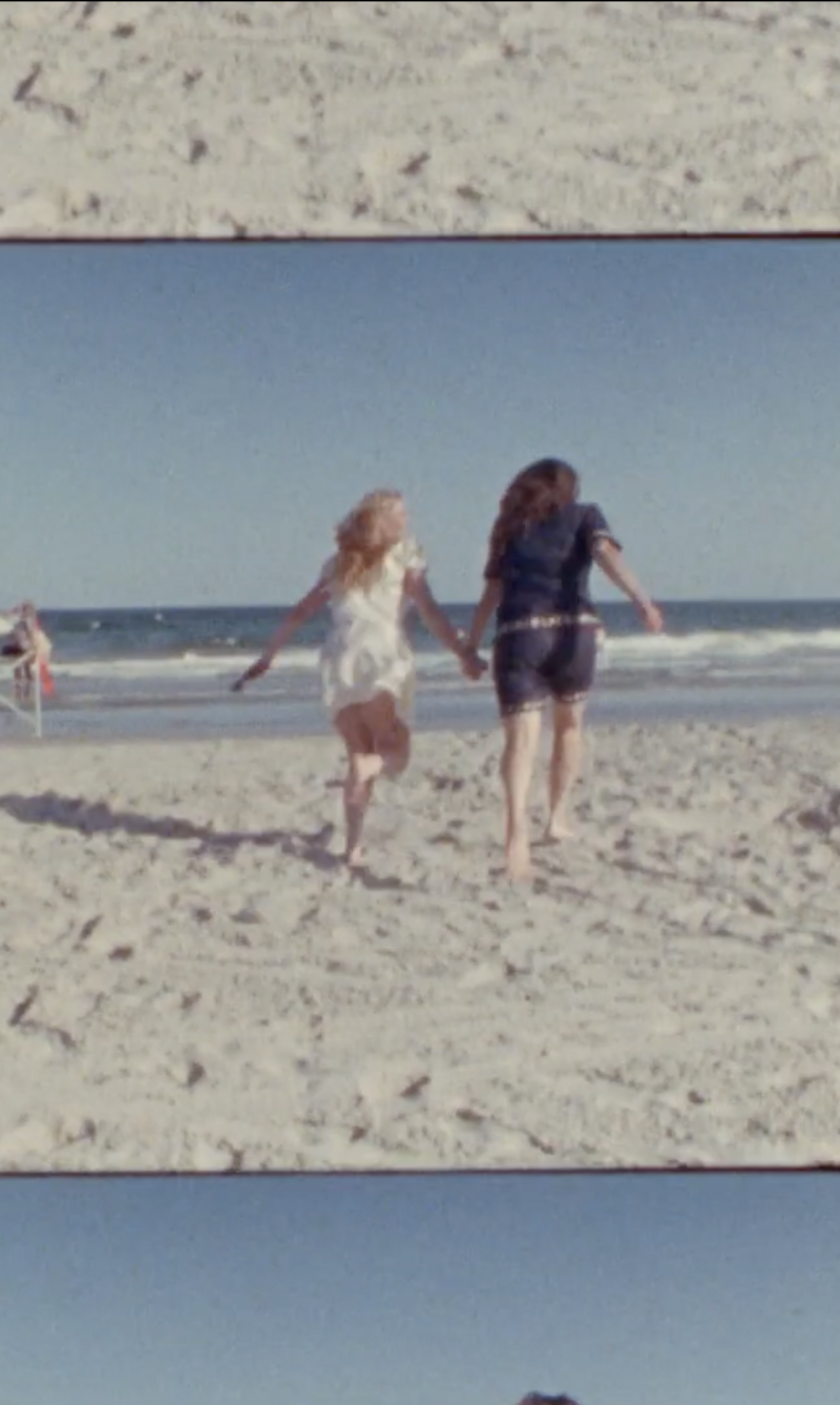 A screenshot from a Super 8 video of two women holding hands and running on the beach