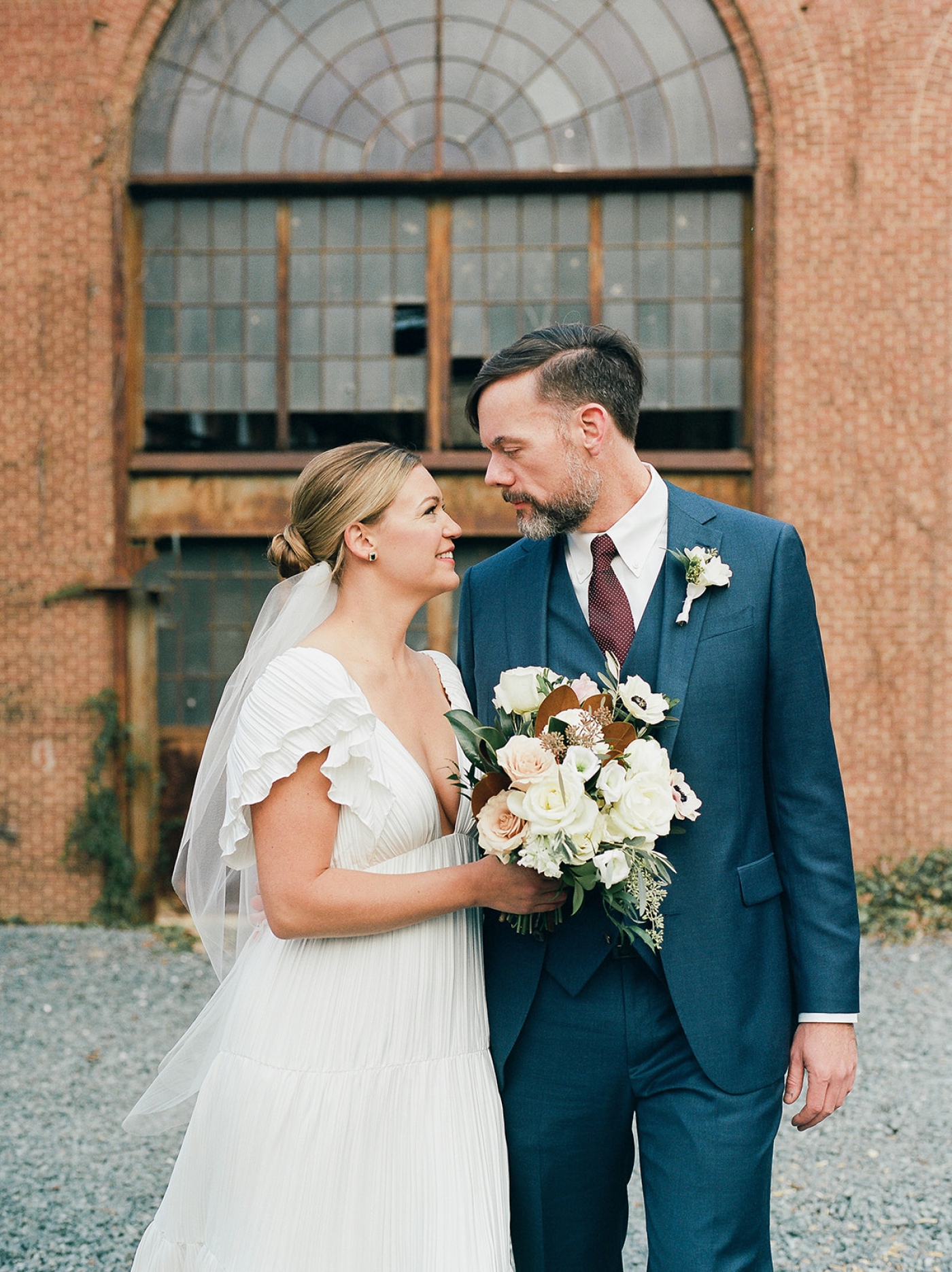 A bride and groom look at each other while standing in front of a large window in a factory in baltimore